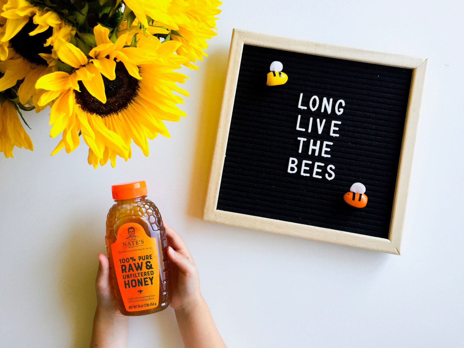 Hand holding Nate’s honey bottle and letterboard saying LONG LIVE THE BEES with sunflowers.