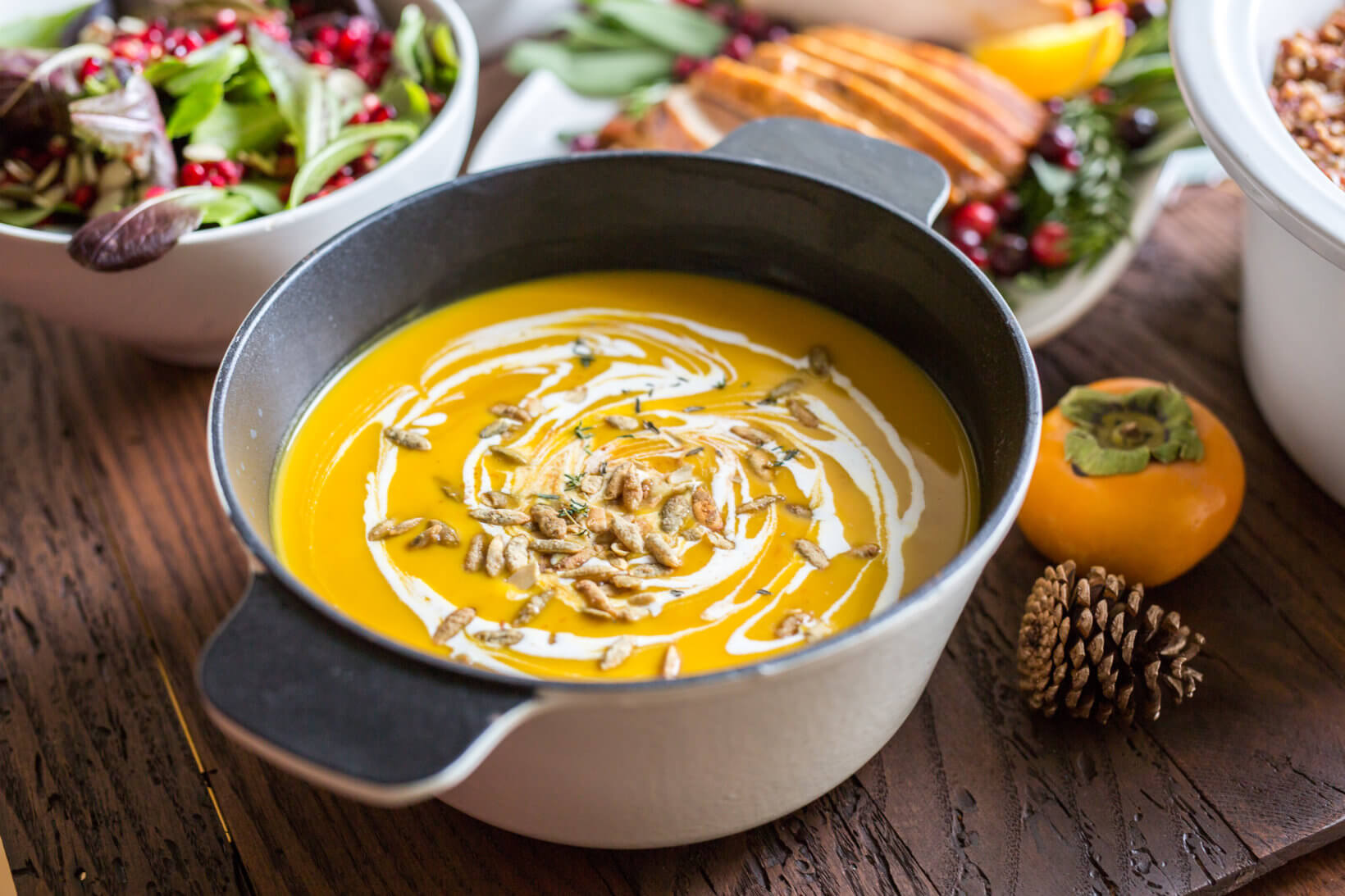 A pot of creamy squash soup made with honey and garnished with seeds and cream, surrounded by autumnal decor and salads.