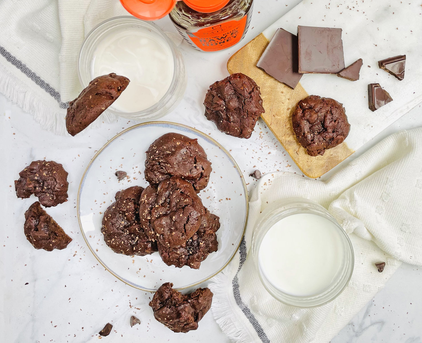 Chocolate cookies on a plate with chunks of chocolate, two glasses of milk, and a Nate’s honey bottle on marble.