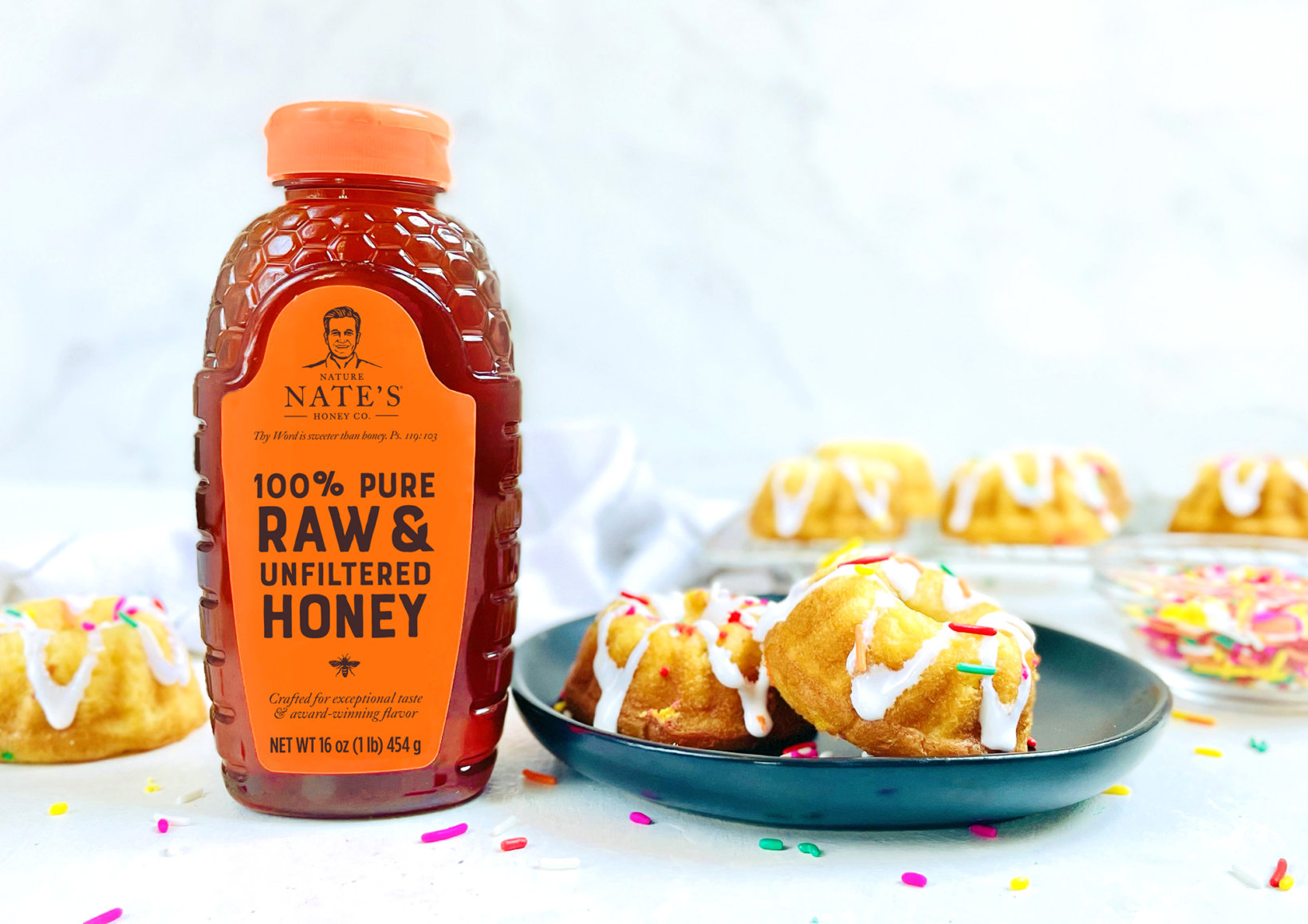 A bottle of Nate's honey with a plate of frosted mini bundt cakes and colorful sprinkles on a marble surface.