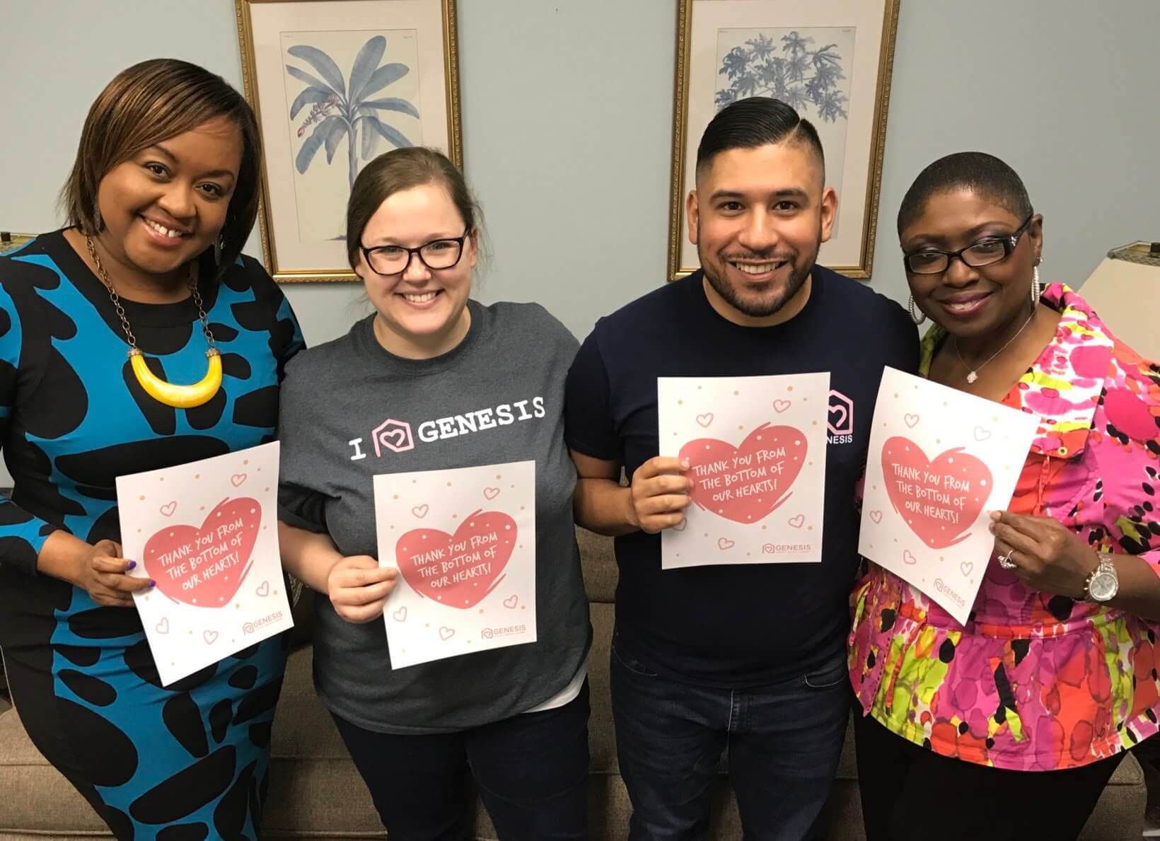 Four people smiling and holding Valentine's Day cards with hearts and messages, with a palm tree picture on the wall.