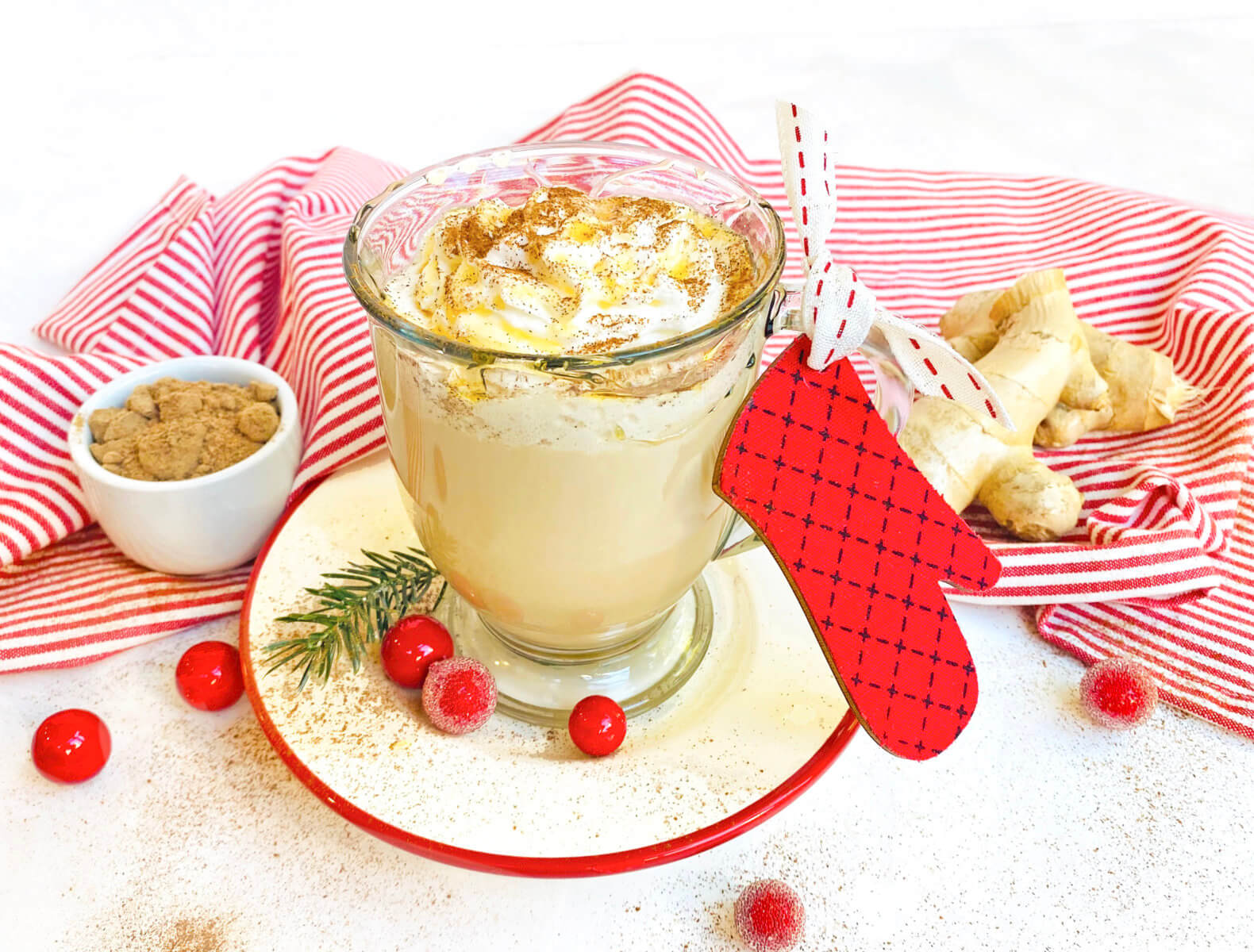 A festive gingerbread latte with whipped cream, a candy cane, and holiday decorations on a red-striped cloth.