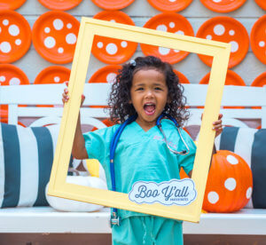 A joyful child in a blue scrub costume holds a yellow frame, with Boo Y'all text, in front of a wall.