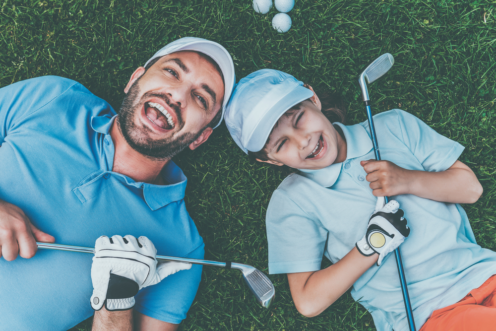 Father and son lying on grass with golf clubs, laughing together.