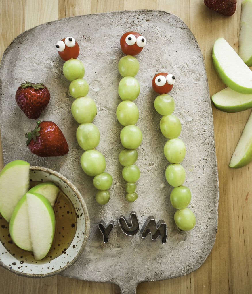 Grape caterpillars with eye decorations on a tray with apple slices, strawberries and yum spelled out with cutters.