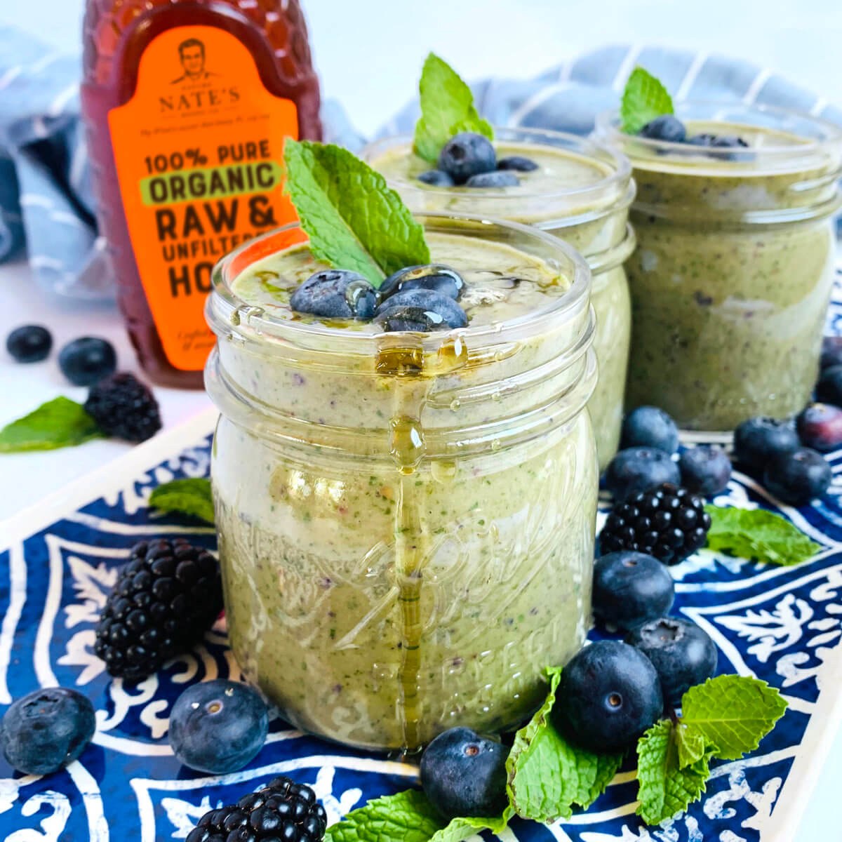 Green smoothie in jars, Nate's organic honey bottle beside blueberries, blackberries and mint on blue patterned cloth.