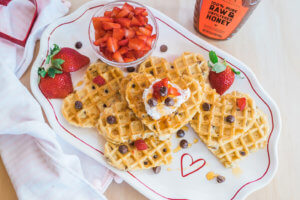 Heart-shaped waffles with a cream, fresh strawberries and chocolate chips served next to a bottle of Nate's honey.