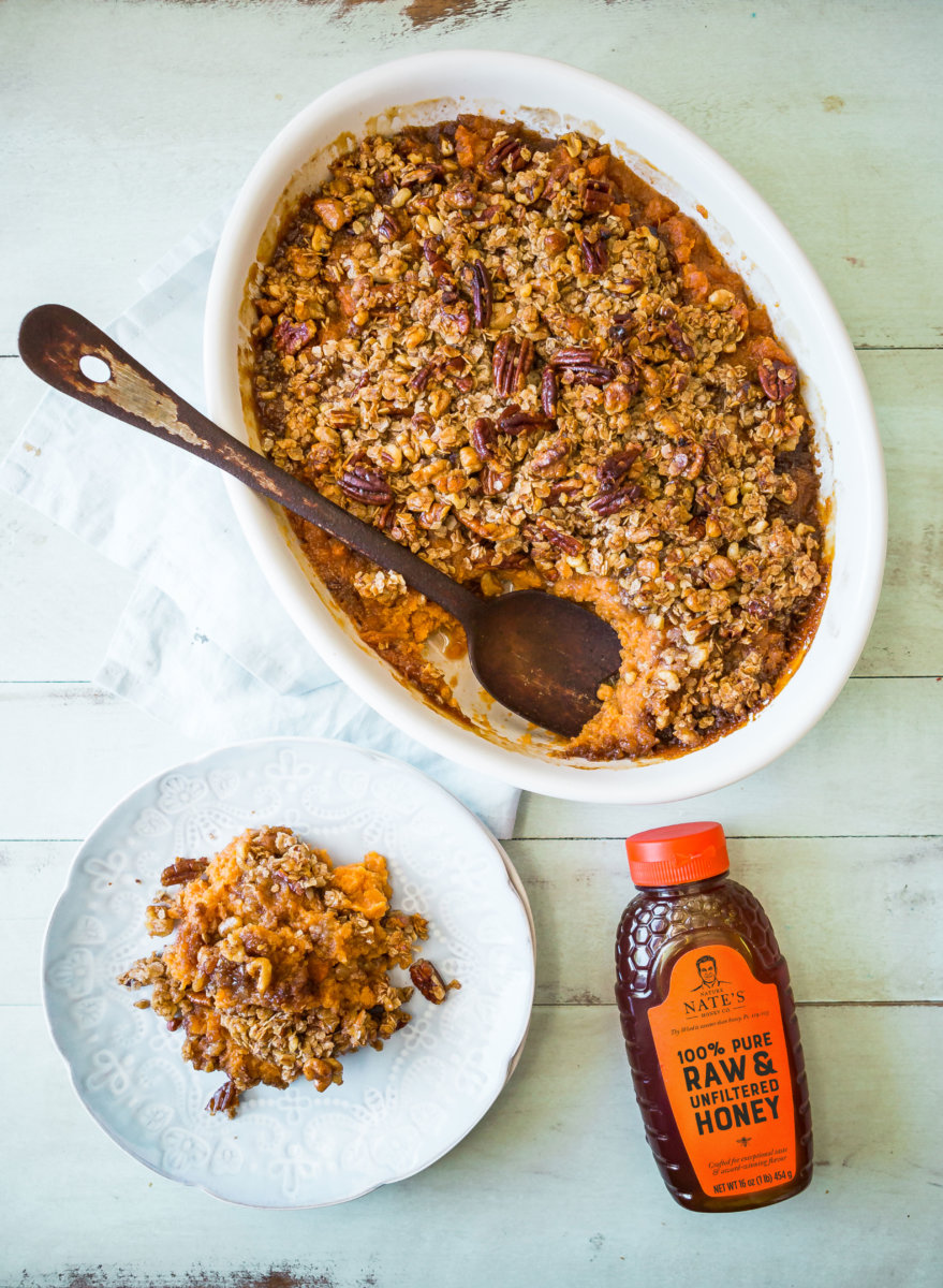 Baked sweet potatoes with crunchy topping in a dish with a serving on a plate and Nate's honey bottle aside.