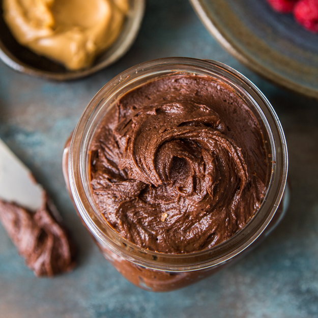 A jar filled with chocolate spread on a textured blue surface, with a butter knife and raspberries nearby.