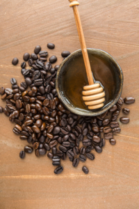 Honey with dipper in a bowl with coffee beans on a wooden surface.