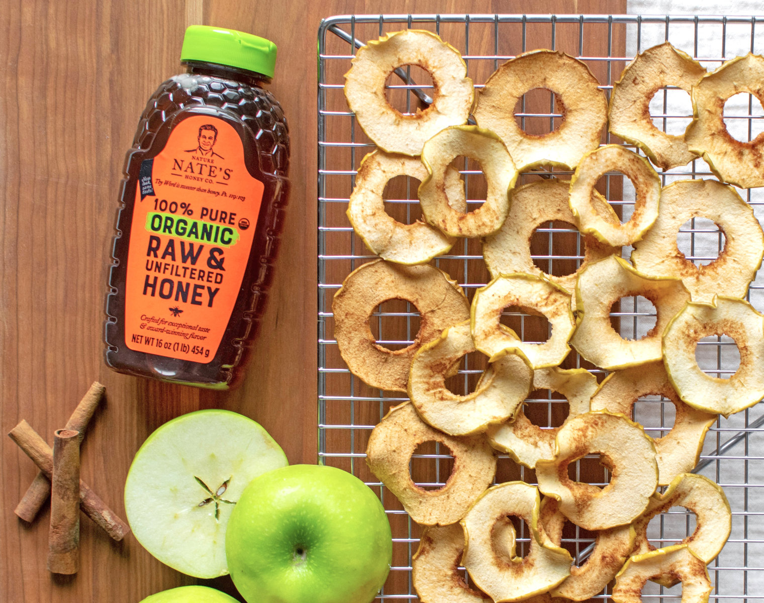 Apple slices dried on a cooling rack with a bottle of Nate’s organic honey and fresh green apples.