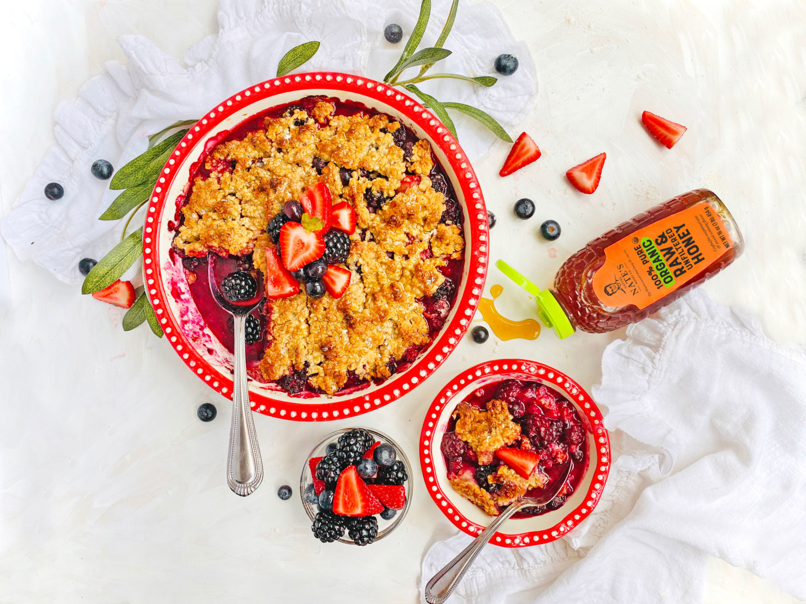 Berry crumble in red dishes with a bottle of Nate’s organic honey and fresh berries on a white cloth.
