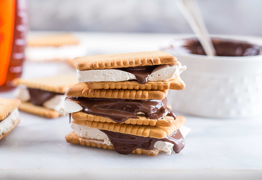 Stack of graham crackers with marshmallow and melted chocolate oozing out, with a bowl of chocolate in the background.