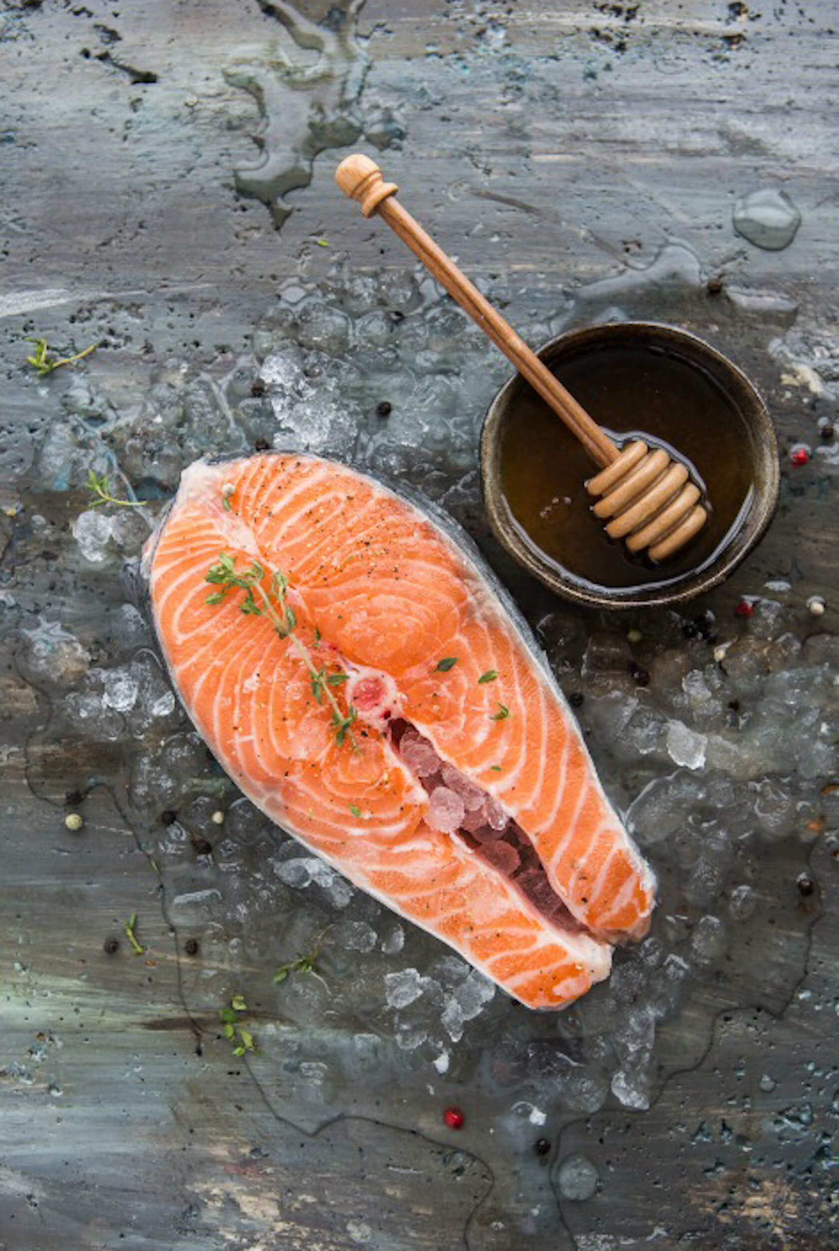 Fresh salmon fillet with herbs and honey with dipper in a bowl on an icy metal surface.