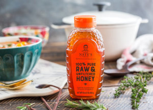 Nate's honey bottle on a rustic table with soup pot and fresh herbs in soft focus background.