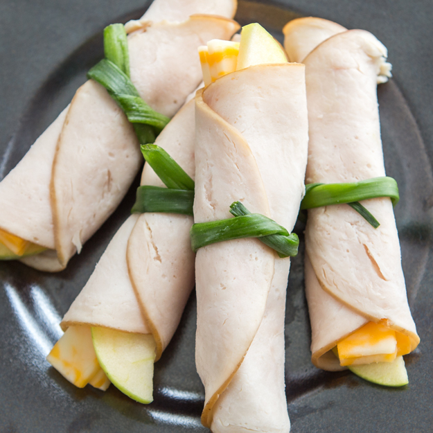Turkey and cheese roll-ups with apple slices on a dark plate.