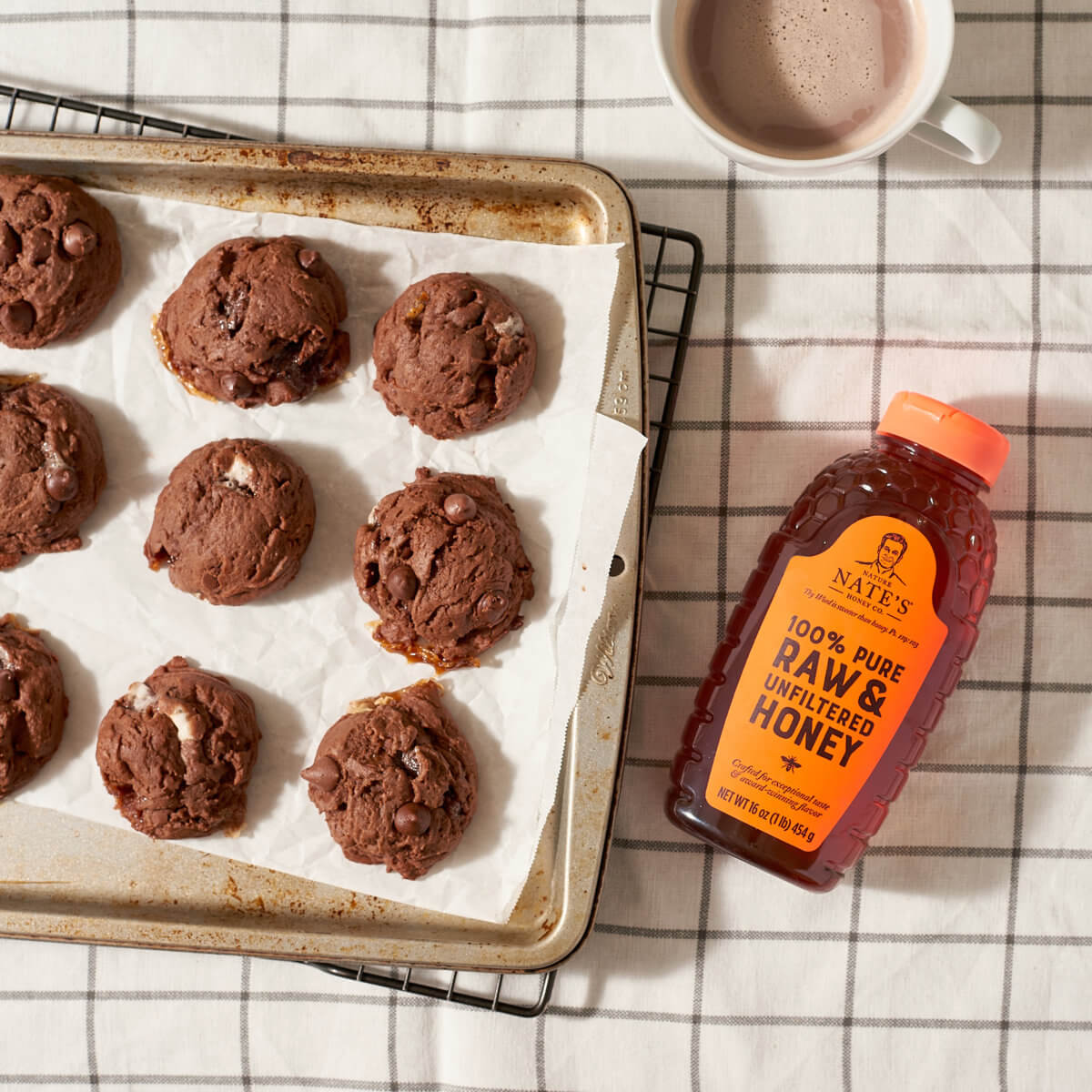 Chocolate cookies on a baking sheet with a bottle of Nate’s honey and a cup of hot cocoa on a checked cloth.