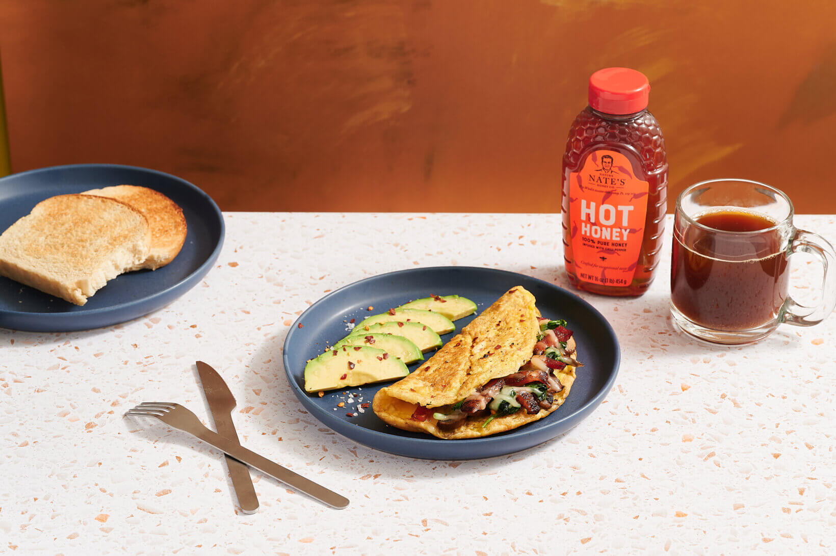 Breakfast set with bacon omelette, avocado, toast, coffee and Nate’s hot honey on an orange surface.