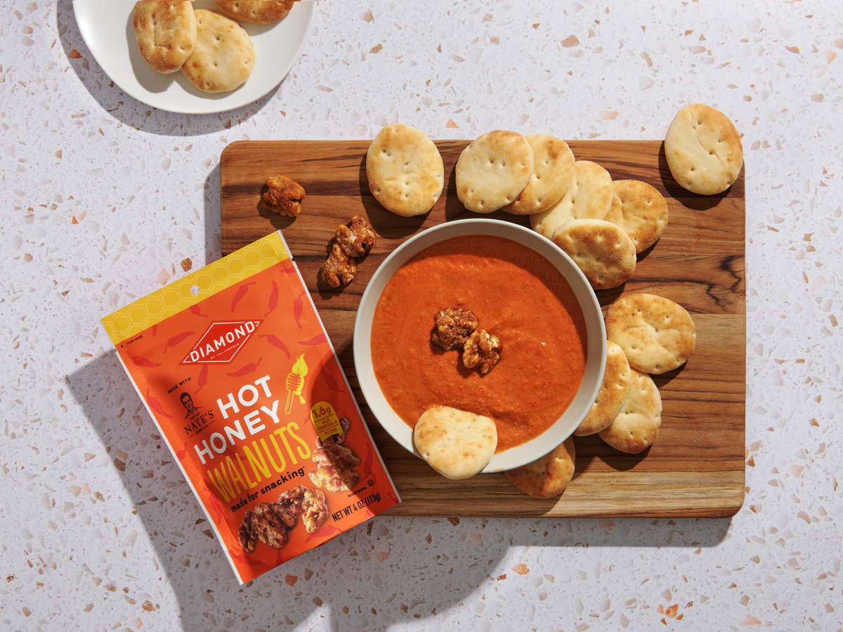 A bowl of red pepper dip on a wooden board, surrounded by bread and a packet of Diamond hot honey walnuts.