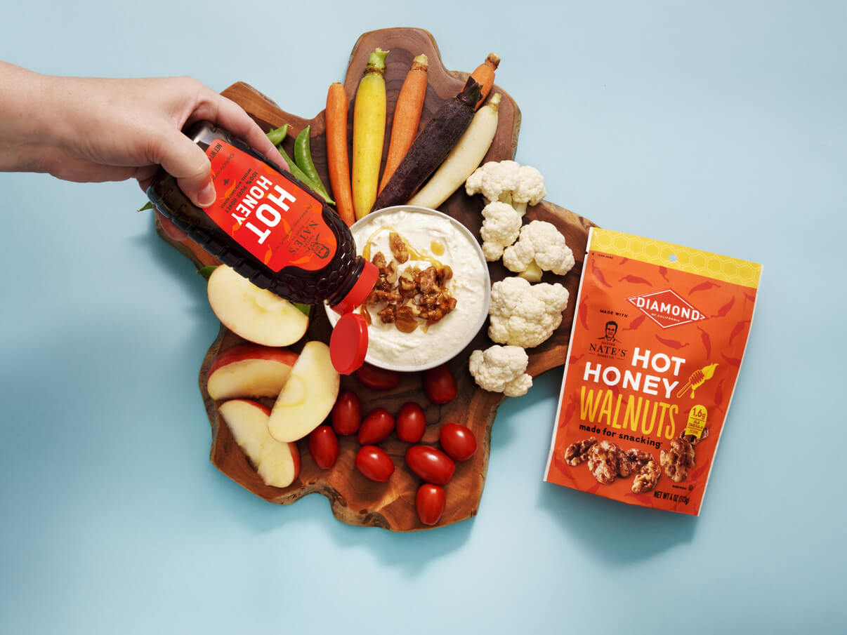 A hand pouring Nate’s hot honey onto whipped feta cheese with veggies, fruits and a Diamond hot honey walnuts packet.