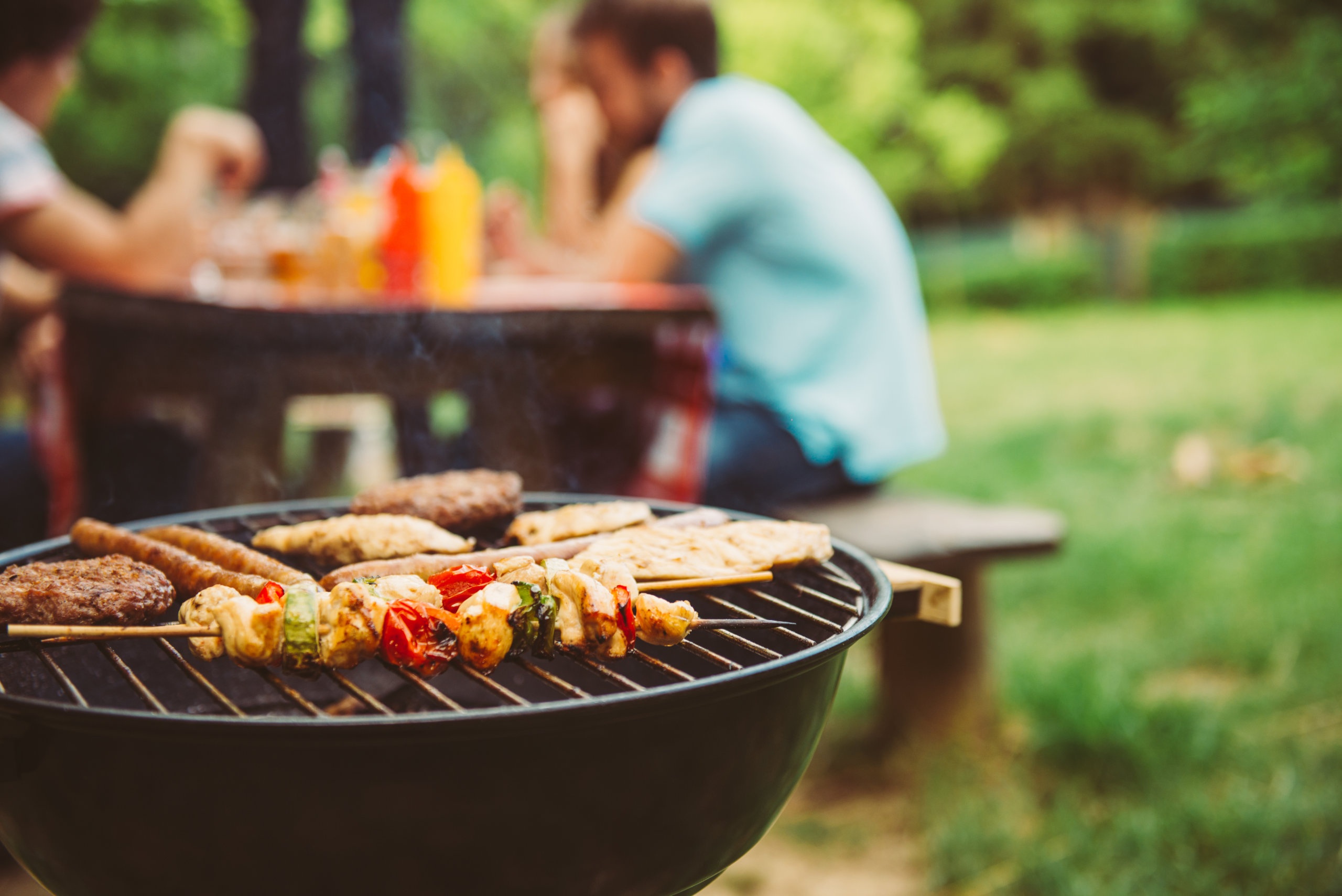 Barbecue grill with food in a park, blurred people in the background.
