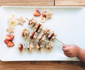 Child's hand picks skewer with banana, strawberry, sandwich bites, and star-shaped bread on white platter.