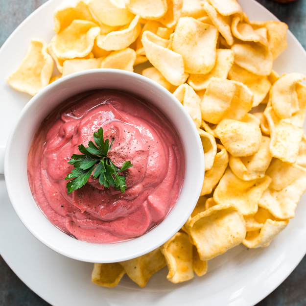 Beet hummus in a bowl with chips and parsley garnish on a white plate.