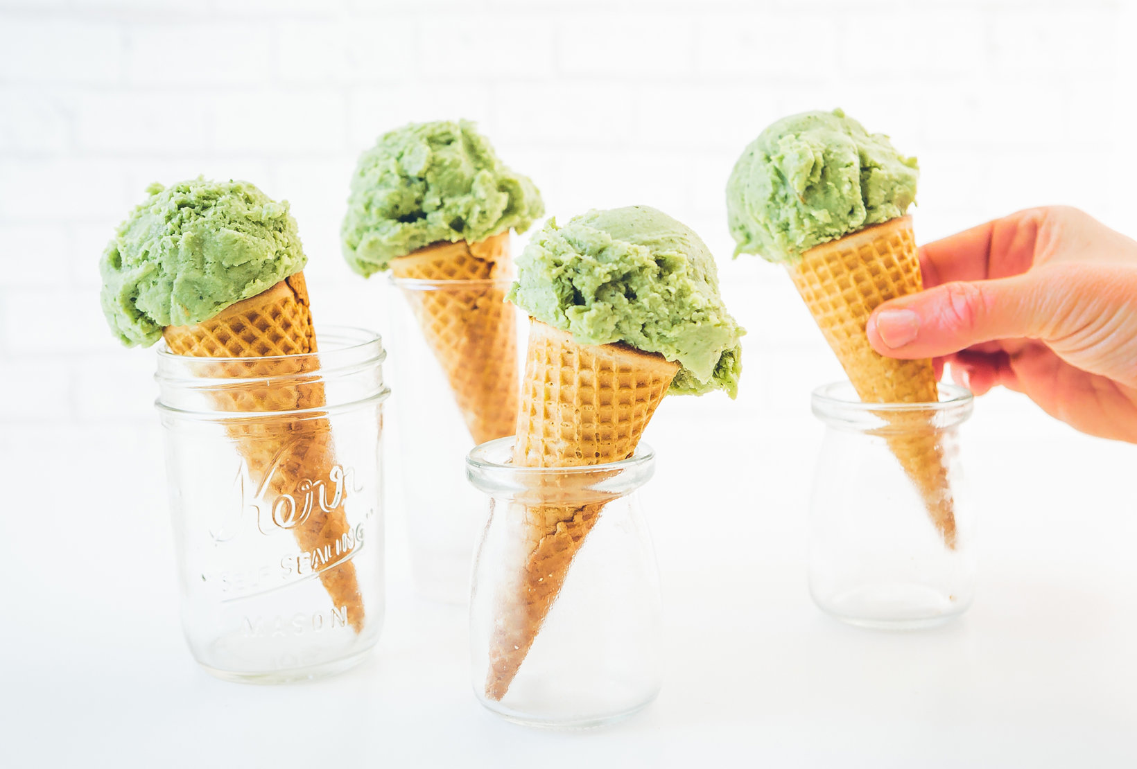 Hand holding a cone of avocado ice cream with more cones in jars on a white table.
