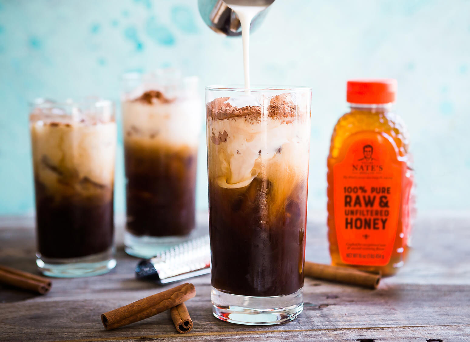 Pouring cream into iced coffee, with more glasses, cinnamon and a bottle of Nate's honey.