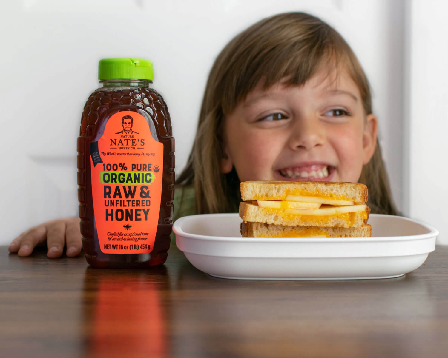 A smiling girl peeking behind a apple, cheese and honey sandwich beside a bottle of Nate’s organic honey on a table.