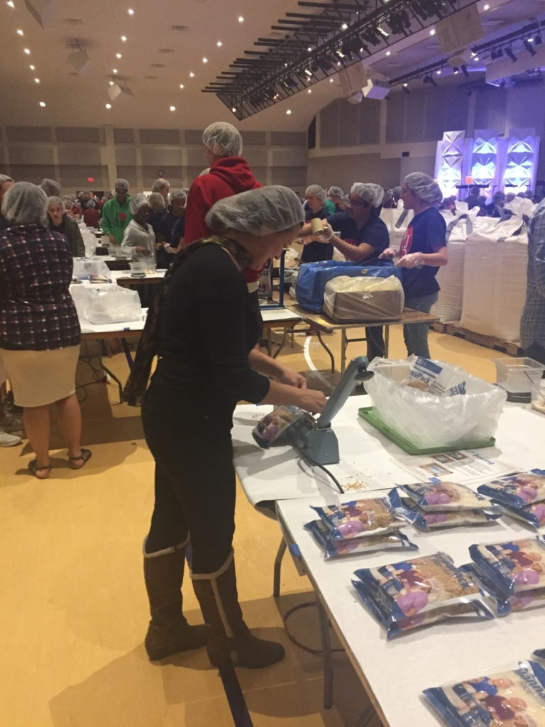 A person in a hairnet is focused on sealing food packages at an event, with other volunteers working in the background.