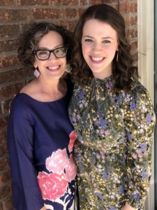 Two smiling women, one in glasses and a floral dress, stand side by side, sharing a joyful moment.