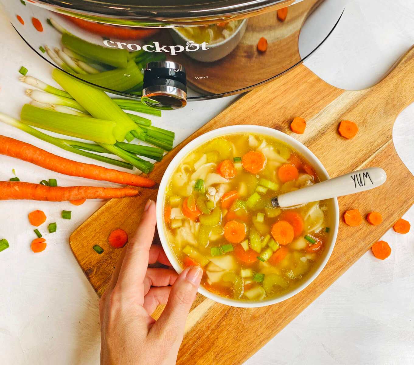  Hand holding a bowl of chicken noodle soup made with Nate’s honey near a slow cooker.