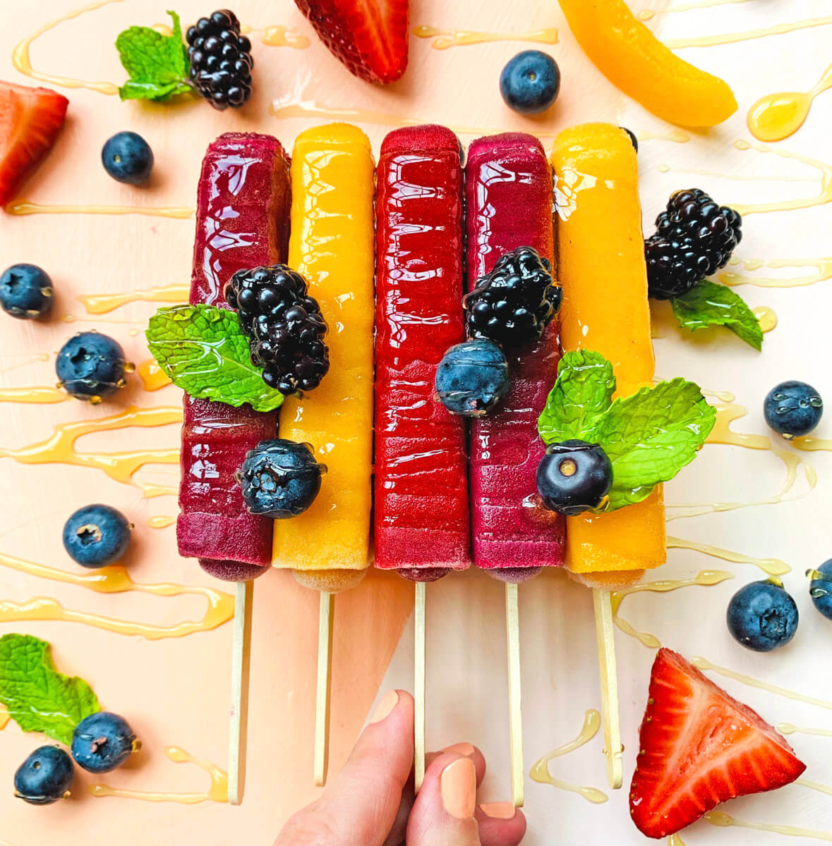 Hand holding fruit popsicles with fresh berries and mint on a drizzled honey background.