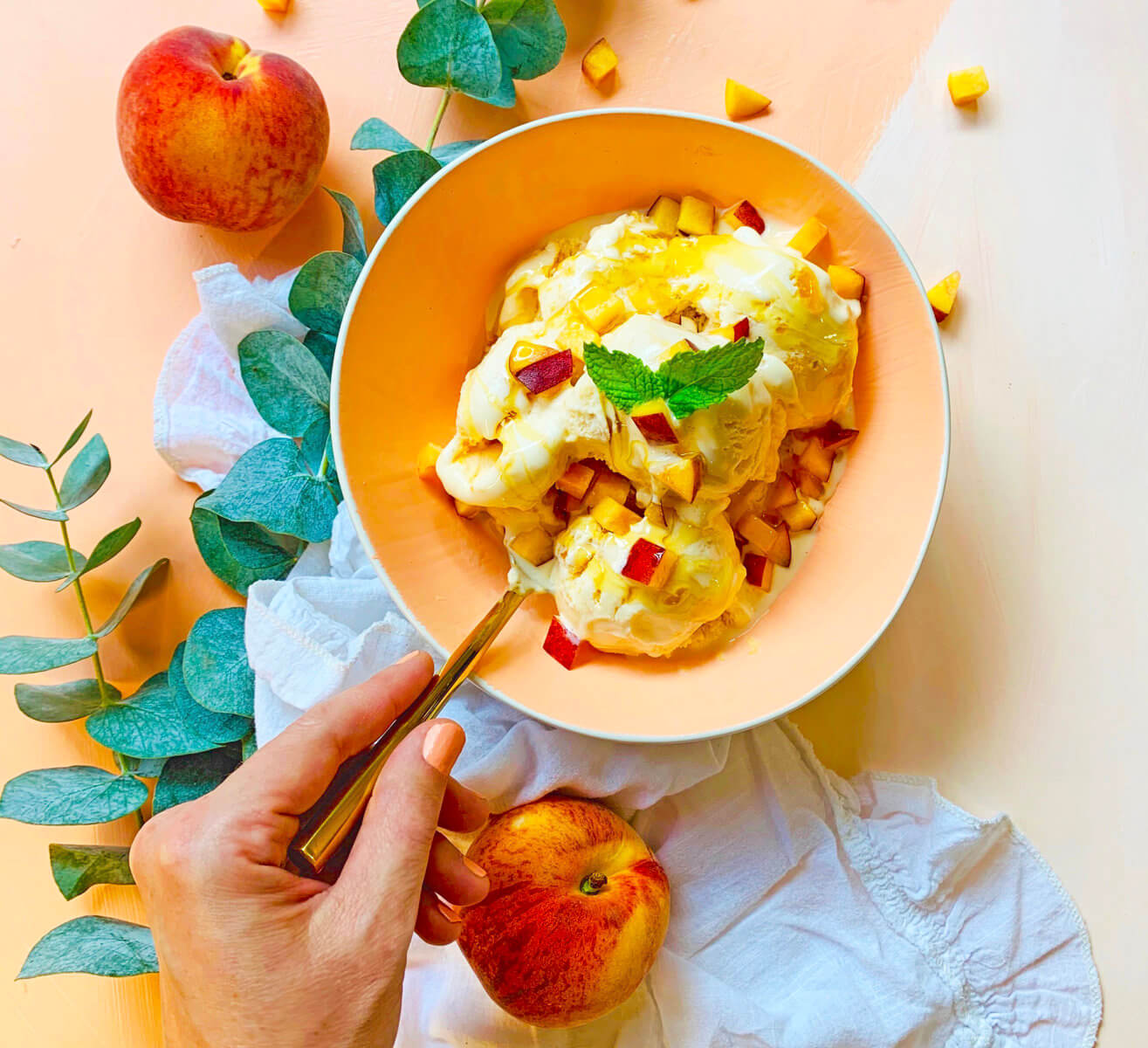 Hand holding a bowl of peaches and honey ice cream with leaves, whole peaches  and a cloth on a peach background.