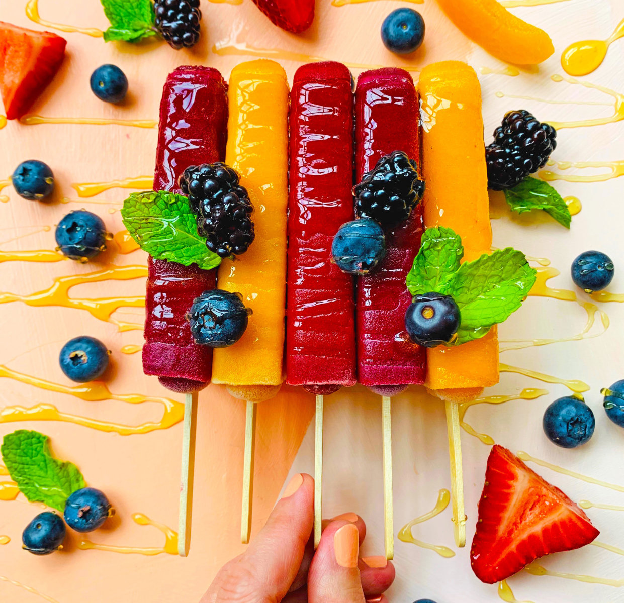 A hand holding colorful fruit popsicles drizzled with honey, garnished with fresh blueberries, mint and strawberries.