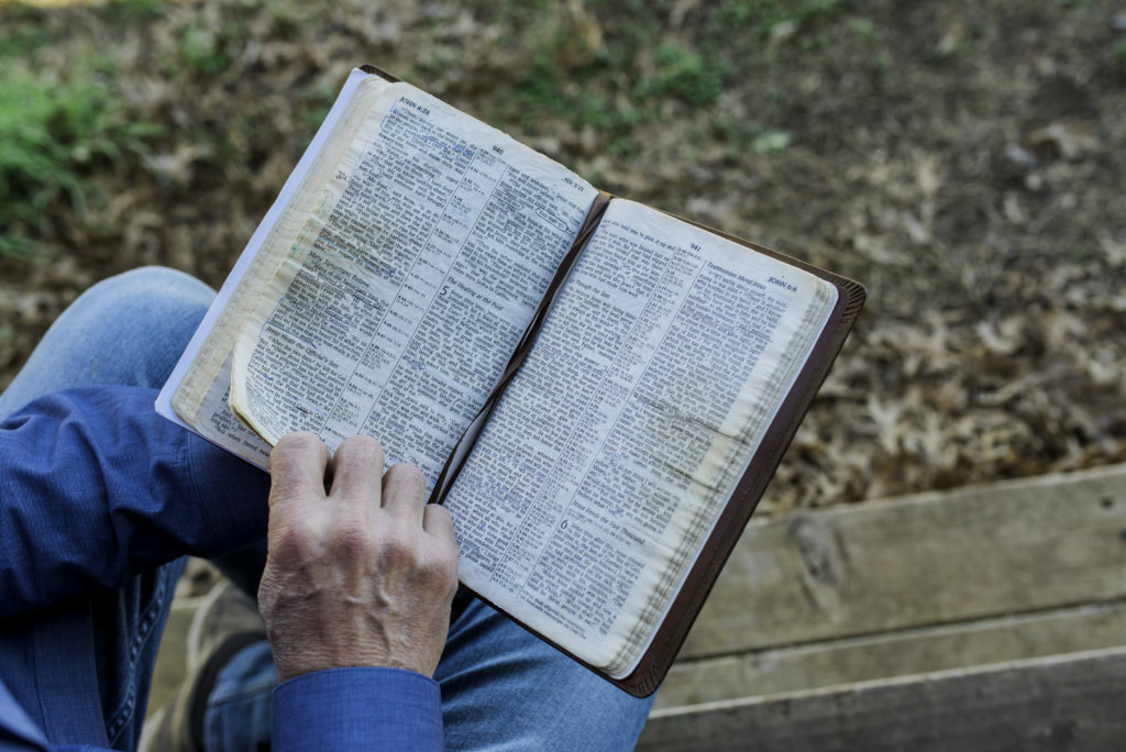 A person in blue jeans reads an open Bible, their hand resting on the pages.