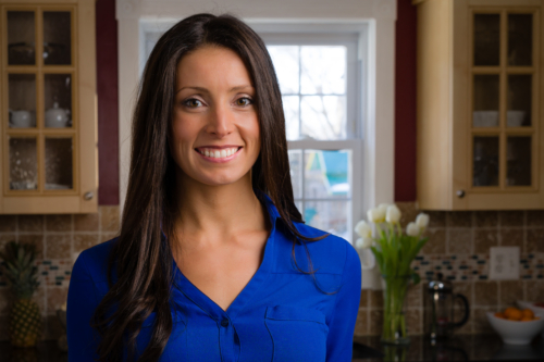 Woman in blue blouse smiling in a kitchen with tulips and fruit bowl in the background.