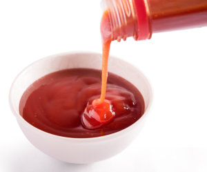 Ketchup pouring from a bottle into a small bowl on a white background.