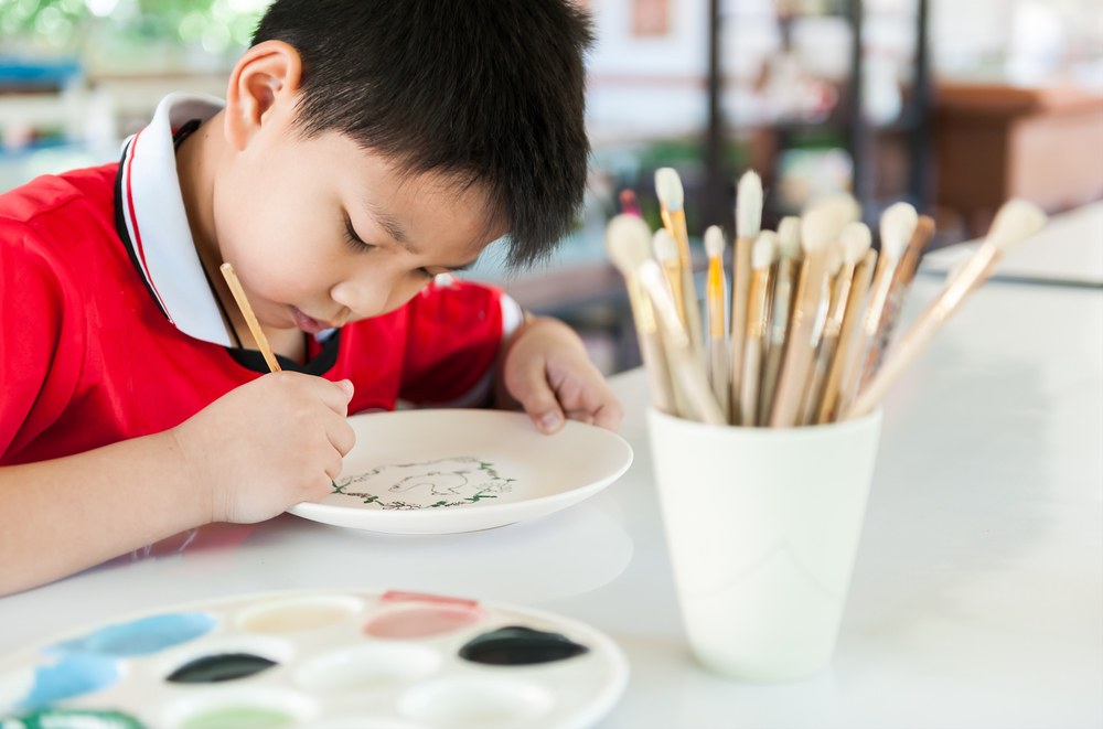 Focused boy painting on a plate with brushes and colors on a table.