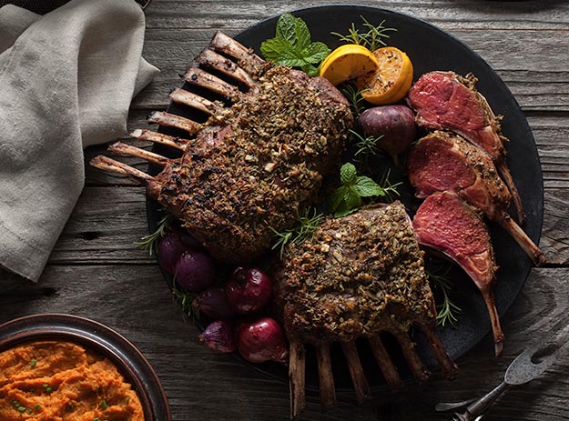 Herb-crusted racks of lamb with roasted vegetables and a napkin on a wooden table.