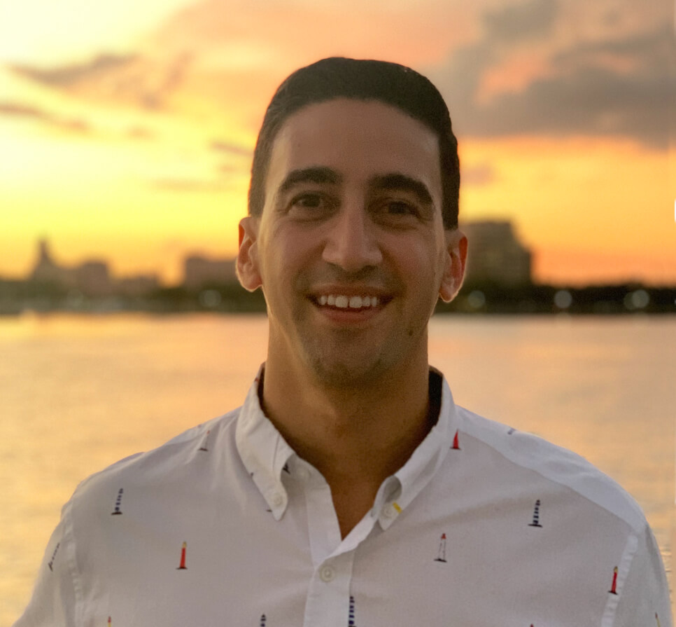 A smiling man in a white shirt with sailboat prints, outdoors during sunset with a waterfront view in the background.