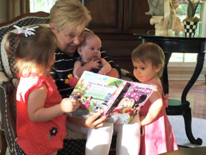 A happy moment with a grandmother and three young children, one a baby, engaged in reading a colorful book together.