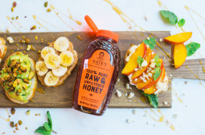Avocado toast, peach toast and sliced banana toast, all drizzled with honey on board with a bottle of Nate's honey.