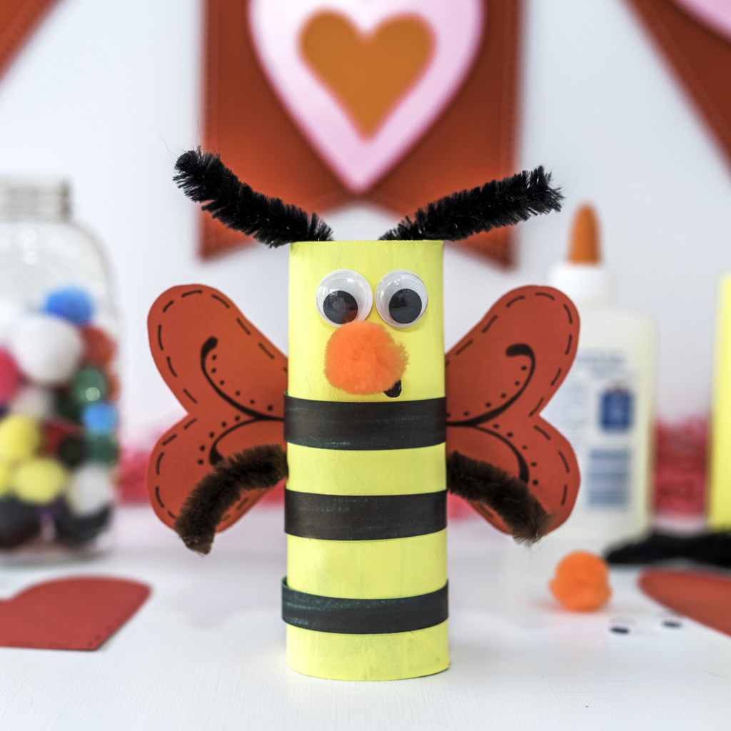 Handcrafted bee with heart wings, googly eyes, on a crafting table.