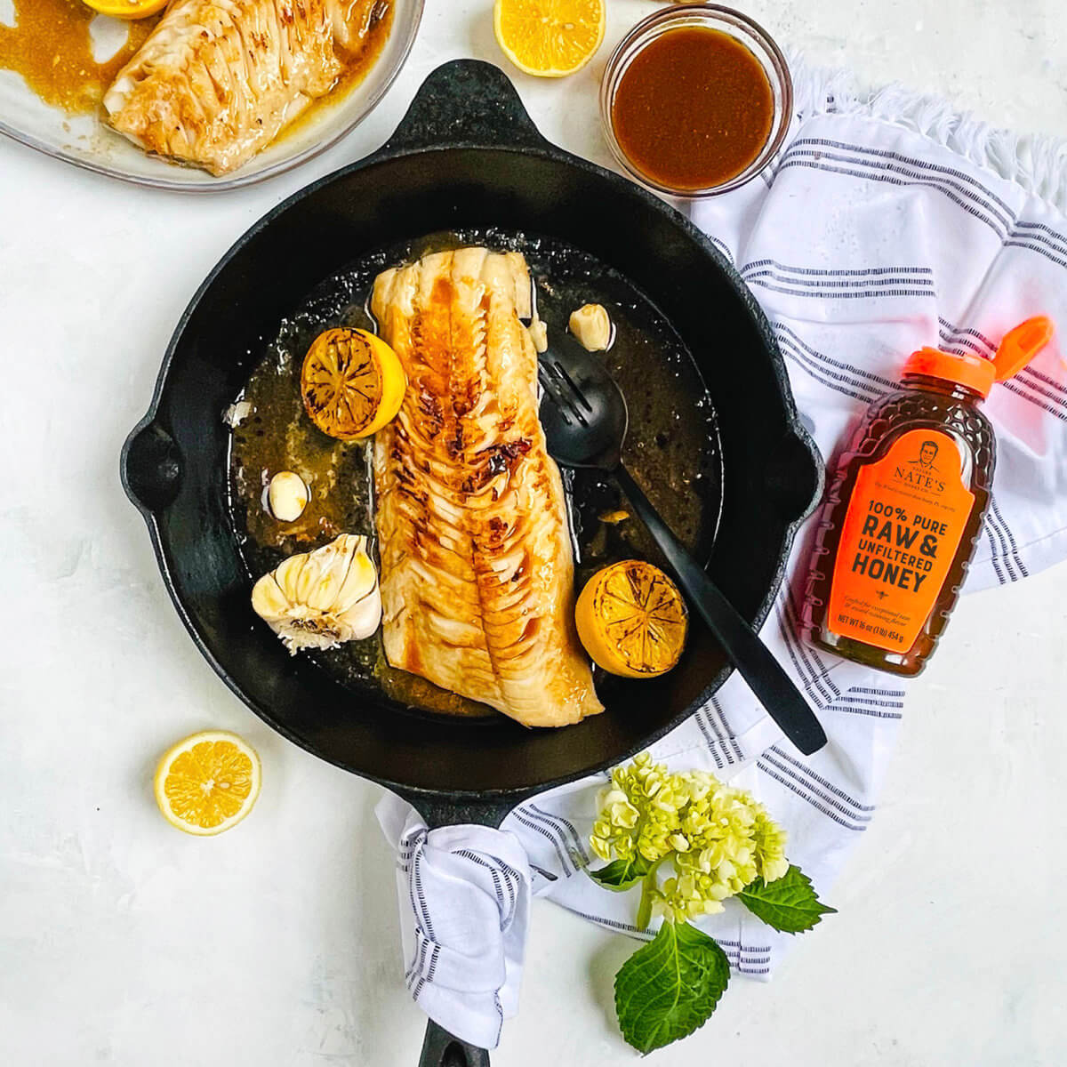 Pan-seared fish fillet in a skillet with lemon slices and garlic cloves, with bottle of Nate's honey and a sauce bowl.