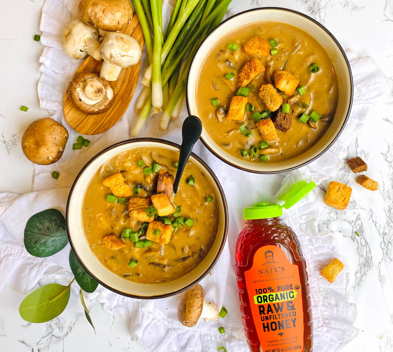 Two bowls of creamy mushroom soup with croutons, beside fresh ingredients and a bottle of Nate’s organic honey.
