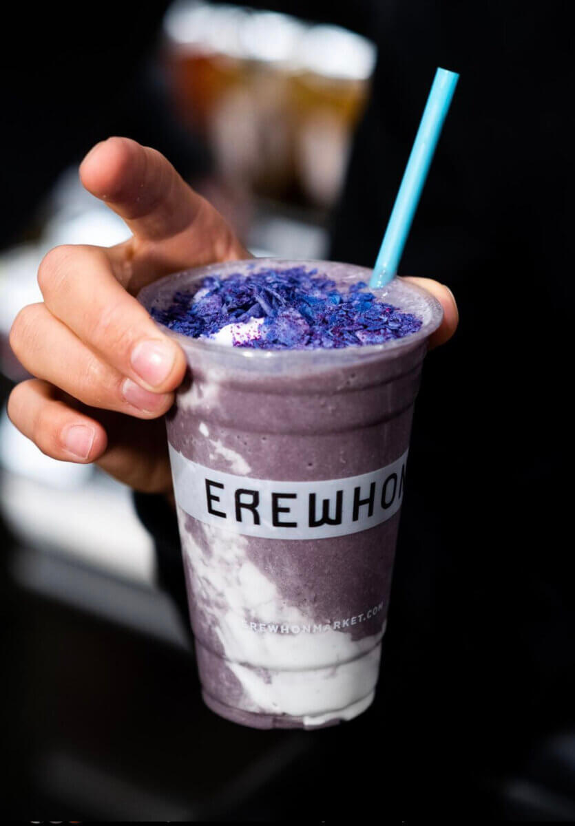 A hand holding a cup with the label EREWHON filled with a layered purple smoothie made with honey.