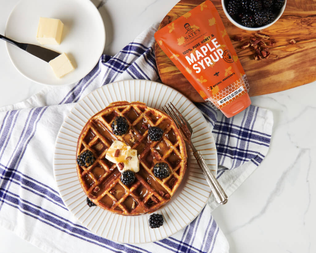 A waffle with butter and blackberries on a plate, Nate's maple syrup and a bowl of blackberries on a marble surface. 