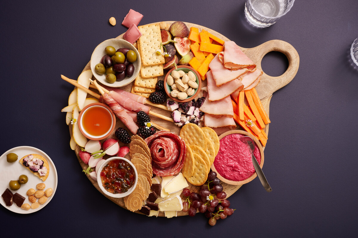 An assortment of meats, cheeses, crackers, nuts and fruits on a wooden charcuterie board with dips and honey in bowls.